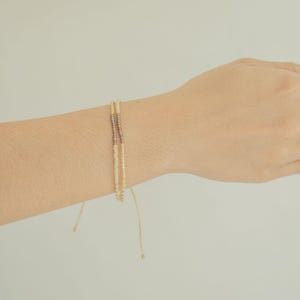 May include: A delicate double-strand bracelet featuring small cream-colored beads and a short section of metallic bronze-toned beads. The bracelet is designed with thin, adjustable cord ends that hang loosely from the main beaded structure.