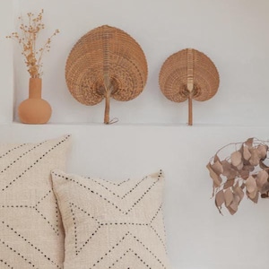 May include: Two woven palm leaf hand fans with a natural tan finish, positioned above two square off-white throw pillows featuring a black stitched chevron pattern. A terracotta vase with dried flowers sits to the left of the fans.