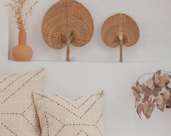 May include: Two woven palm leaf hand fans with a natural tan finish, positioned above two square off-white throw pillows featuring a black stitched chevron pattern. A terracotta vase with dried flowers sits to the left of the fans.