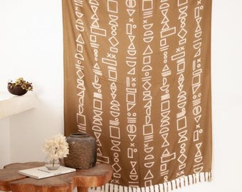 May include: A rectangular brown wall hanging featuring a repeating pattern of white geometric shapes including triangles, circles, squares, and lines. The fabric has a fringed bottom edge and is suspended from the top with small loops.