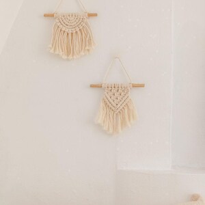 Peut inclure: Deux décorations murales en macramé blanc cassé suspendues à des tiges en bois clair avec des cordons de suspension. La pièce supérieure présente un motif de nœuds en demi-cercle, tandis que la pièce inférieure a un motif triangulaire.