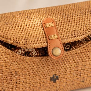 A close-up image of a woven straw bag with a leather strap and a metal button.