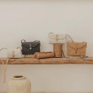 Op de afbeelding: Een collectie rechthoekige en cilindervormige gevlochten tassen in wit, zwart en natuurlijke bruintinten. Elke tas heeft een klepsluiting met een lichtbruine leren riem, een metalen drukknoop en een dunne bijpassende schouderriem om te dragen.