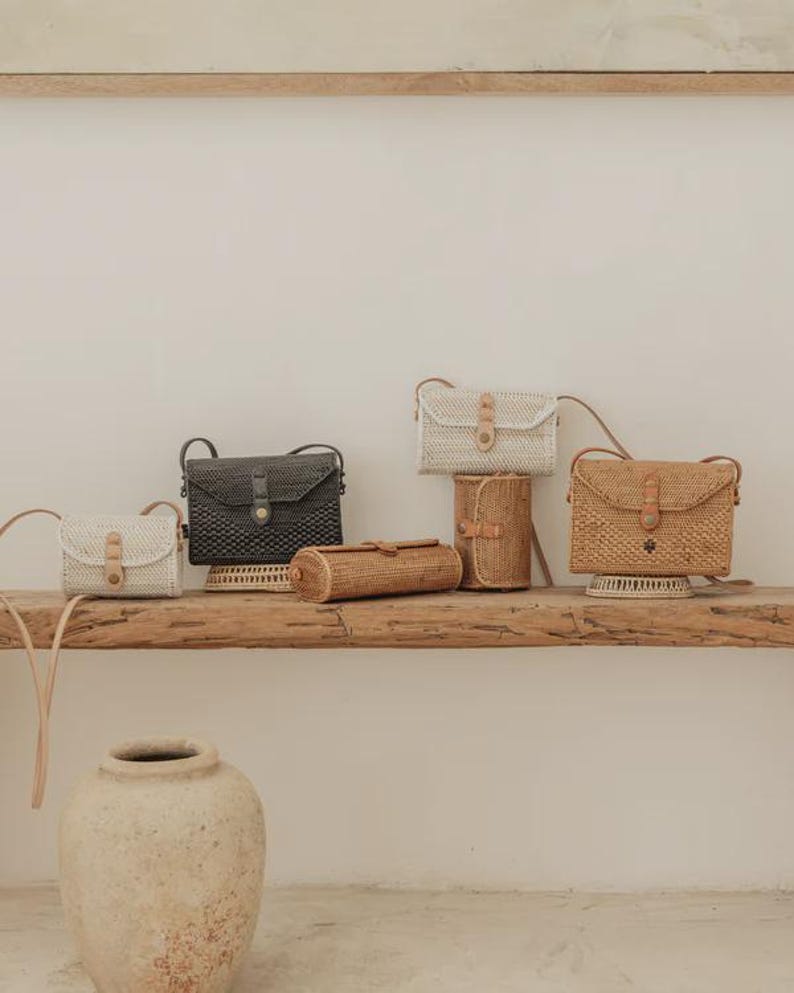 Op de afbeelding: Een collectie rechthoekige en cilindervormige gevlochten tassen in wit, zwart en natuurlijke bruintinten. Elke tas heeft een klepsluiting met een lichtbruine leren riem, een metalen drukknoop en een dunne bijpassende schouderriem om te dragen.