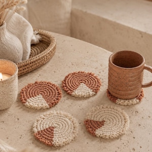May include: A set of four round macrame coasters featuring a spiral pattern in terracotta and cream colors. Each coaster has a fringe edge and a contrasting triangular segment. One coaster is placed under a speckled brown ceramic mug.