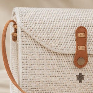 Op de afbeelding: Een rechthoekige, gebroken witte geweven schoudertas met een beige leren riem en een bijpassende leren sluiting die wordt vastgezet met een metalen drukknoop. De tas heeft een gestructureerd weefpatroon en een klein donkergrijs kruis-embleem.