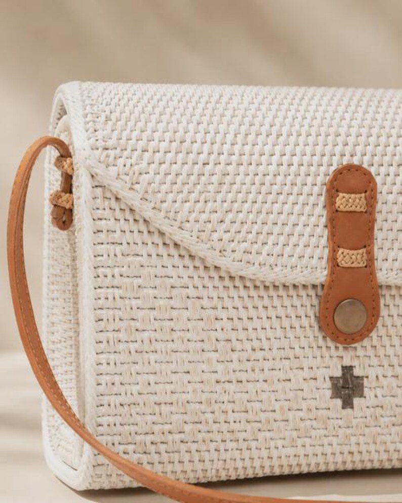 Op de afbeelding: Een rechthoekige, gebroken witte geweven schoudertas met een beige leren riem en een bijpassende leren sluiting die wordt vastgezet met een metalen drukknoop. De tas heeft een gestructureerd weefpatroon en een klein donkergrijs kruis-embleem.