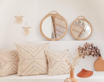 May include: Two round rattan-framed mirrors, three square beige throw pillows with black stitched geometric patterns, two small macrame wall hangings, a brown woven visor, two terracotta-colored vases, and a dried plant arrangement.