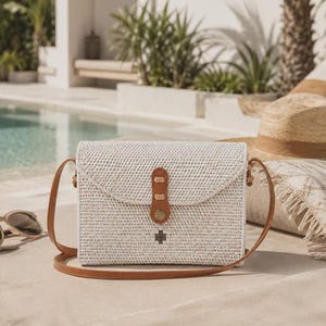 Op de afbeelding: Een rechthoekige crossbodytas van gevlochten wit rotan met een bruine leren schouderband en een bijpassende leren sluiting met een metalen drukknoop. De tas heeft een klein donker metalen kruisvormig embleem op de voorflap.