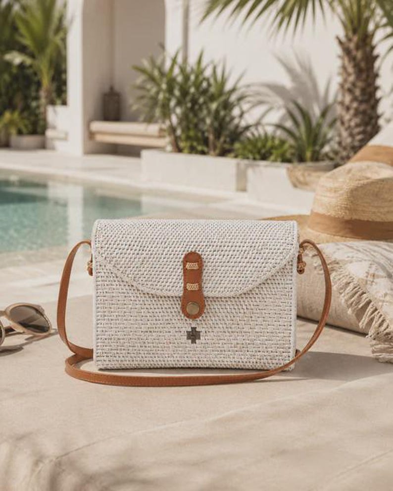 Op de afbeelding: Een rechthoekige crossbodytas van gevlochten wit rotan met een bruine leren schouderband en een bijpassende leren sluiting met een metalen drukknoop. De tas heeft een klein donker metalen kruisvormig embleem op de voorflap.