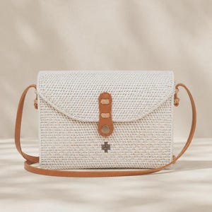 Op de afbeelding: Een rechthoekige geweven schoudertas in off-white met een bruine leren riem en een bijpassende leren sluiting met een metalen drukknoop. Een klein zilverkleurig metalen embleem is gecentreerd op de voorflap van de tas.
