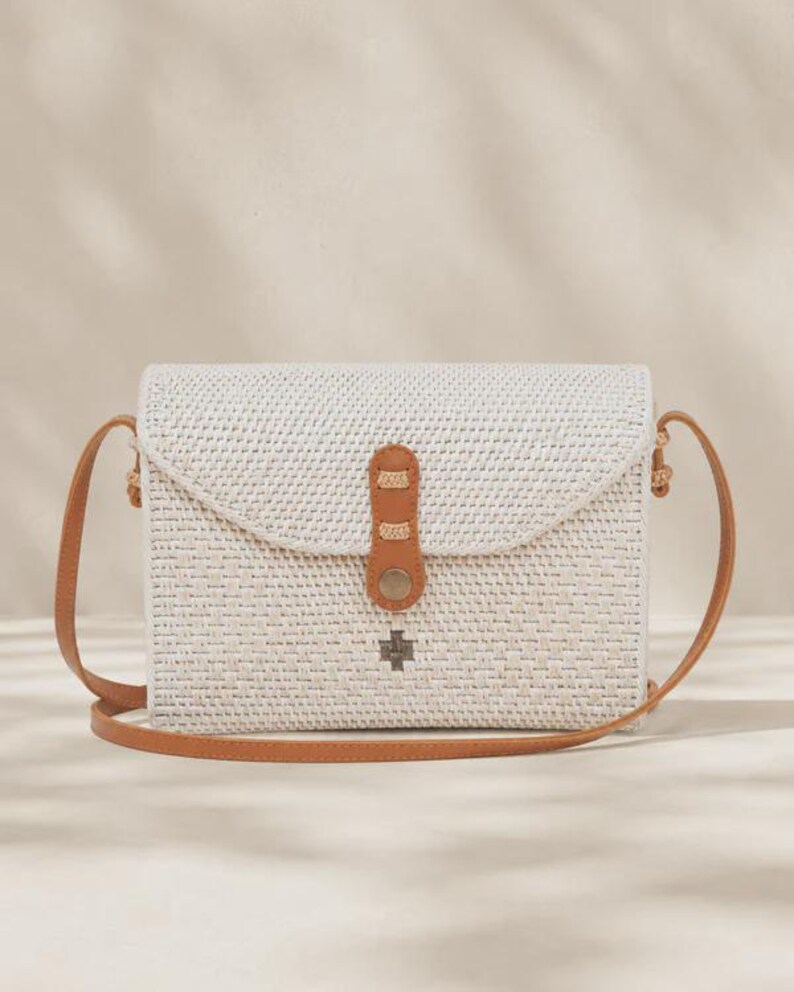 Op de afbeelding: Een rechthoekige geweven schoudertas in off-white met een bruine leren riem en een bijpassende leren sluiting met een metalen drukknoop. Een klein zilverkleurig metalen embleem is gecentreerd op de voorflap van de tas.