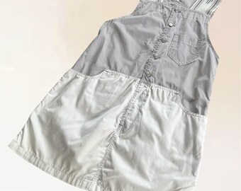 May include: A vintage overall dress featuring a striped top and a solid white skirt. The top has a button-down front, a chest pocket, and striped straps with a small logo. The dress is made of a lightweight fabric.