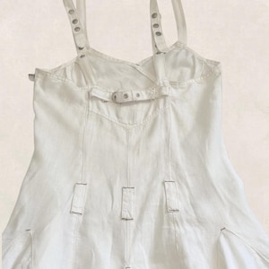 K&ouml;nnte beinhalten: Ein wei&szlig;es &auml;rmelloses Kleid mit verstellbaren Schultertr&auml;gern, die mit silberfarbenen Metall&ouml;sen und Schnallen versehen sind. Das Oberteil verf&uuml;gt &uuml;ber einen horizontalen R&uuml;ckengurt mit Metallschnalle sowie vertikale N&auml;hte mit kleinen Stofflaschen.