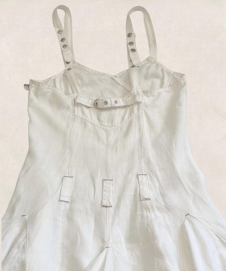 K&ouml;nnte beinhalten: Ein wei&szlig;es &auml;rmelloses Kleid mit verstellbaren Schultertr&auml;gern, die mit silberfarbenen Metall&ouml;sen und Schnallen versehen sind. Das Oberteil verf&uuml;gt &uuml;ber einen horizontalen R&uuml;ckengurt mit Metallschnalle sowie vertikale N&auml;hte mit kleinen Stofflaschen.