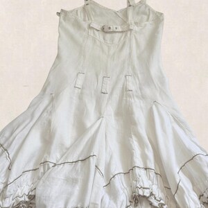 K&ouml;nnte beinhalten: Ein wei&szlig;es &auml;rmelloses Kleid mit verstellbaren Tr&auml;gern und Metallbeschl&auml;gen, einem horizontalen R&uuml;ckengurt mit Schnalle und vertikalen N&auml;hten. Der Rock hat eine ausgestellte Silhouette mit gerafften Stufen und einem d&uuml;nnen braunen Saum.