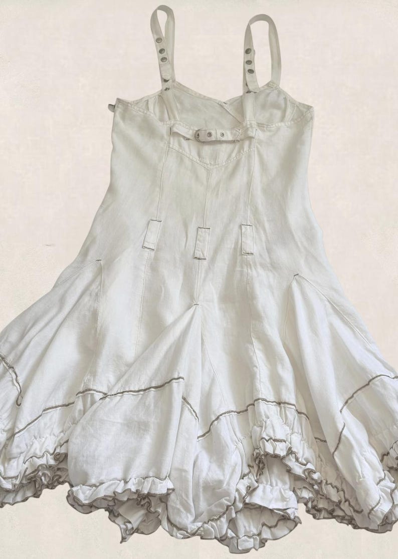 K&ouml;nnte beinhalten: Ein wei&szlig;es &auml;rmelloses Kleid mit verstellbaren Tr&auml;gern und Metallbeschl&auml;gen, einem horizontalen R&uuml;ckengurt mit Schnalle und vertikalen N&auml;hten. Der Rock hat eine ausgestellte Silhouette mit gerafften Stufen und einem d&uuml;nnen braunen Saum.