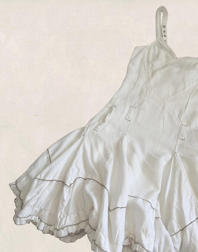 K&ouml;nnte beinhalten: Ein wei&szlig;es &auml;rmelloses Kleid mit einem figurbetonten Oberteil mit sichtbaren N&auml;hten und einem ausgestellten Rock. Der Rock besteht aus mehreren Bahnen und horizontalen Steppn&auml;hten und endet in einem R&uuml;schensaum mit d&uuml;nnen braunen Akzenten.