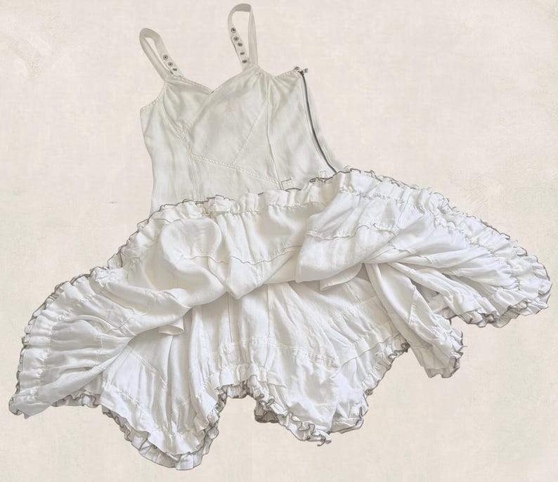K&ouml;nnte beinhalten: Ein wei&szlig;es &auml;rmelloses Kleid mit einem figurbetonten Oberteil, d&uuml;nnen Tr&auml;gern und einem seitlichen Rei&szlig;verschluss. Der Rock ist mit mehreren Lagen geraffter R&uuml;schen gestaltet, was eine volumin&ouml;se, ausgestellte Silhouette mit strukturierter Optik erzeugt.