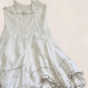 K&ouml;nnte beinhalten: Ein wei&szlig;es &auml;rmelloses Kleid mit verstellbaren Schultertr&auml;gern mit Metallschnallen, einem horizontalen R&uuml;ckengurt mit Schnalle, vertikalen Nahtdetails und einem ausgestellten Rock mit mehreren Stufen von R&uuml;schen und d&uuml;nnen braunen Kontrastn&auml;hten.