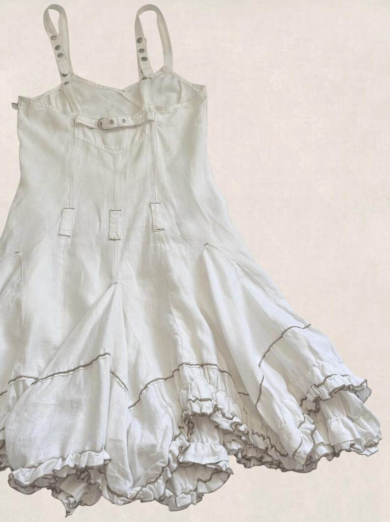K&ouml;nnte beinhalten: Ein wei&szlig;es &auml;rmelloses Kleid mit verstellbaren Schultertr&auml;gern mit Metallschnallen, einem horizontalen R&uuml;ckengurt mit Schnalle, vertikalen Nahtdetails und einem ausgestellten Rock mit mehreren Stufen von R&uuml;schen und d&uuml;nnen braunen Kontrastn&auml;hten.