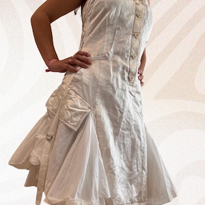 Könnte beinhalten: Ein weißes Kleid mit einem einzigartigen Design. Das Kleid hat ein geknöpftes Oberteil, Taschen und einen mehrlagigen Rock. Der Stoff hat ein strukturiertes Aussehen, und das Kleid hat einen lässigen, aber stilvollen Look. Das Kleid ist ärmellos und hat eine taillierte Taille.