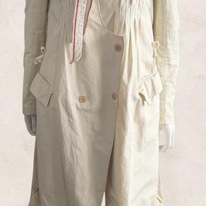 Puede incluir: Abrigo color crema con un diseño único. Presenta un cuello alto, un corpiño plisado y un cierre cruzado con dos botones. El abrigo tiene mangas largas, bolsillos y un acento rojo y blanco. El dobladillo está detallado con encaje.