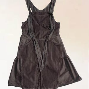 Könnte beinhalten: Dunkelgraues Jeans-Latzkleid mit ausgestelltem Rock und verstellbaren Trägern. Das Kleid hat eine Vordertasche, eine Bindetaille und weiße Nahtdetails. Das Latzkleid ist ein lässiges Kleidungsstück.