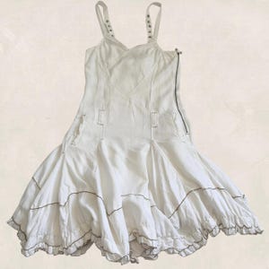 K&ouml;nnte beinhalten: Ein wei&szlig;es &auml;rmelloses Kleid mit einem figurbetonten Oberteil, d&uuml;nnen Tr&auml;gern, einem seitlichen Rei&szlig;verschluss und einem ausgestellten Rock aus mehreren Bahnen. Der Rock hat horizontale N&auml;hte mit sichtbaren Stichen und einen R&uuml;schensaum.