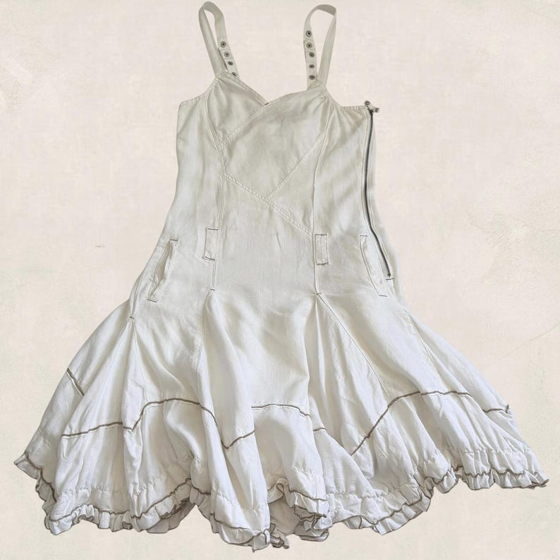 K&ouml;nnte beinhalten: Ein wei&szlig;es &auml;rmelloses Kleid mit einem figurbetonten Oberteil, d&uuml;nnen Tr&auml;gern, einem seitlichen Rei&szlig;verschluss und einem ausgestellten Rock aus mehreren Bahnen. Der Rock hat horizontale N&auml;hte mit sichtbaren Stichen und einen R&uuml;schensaum.