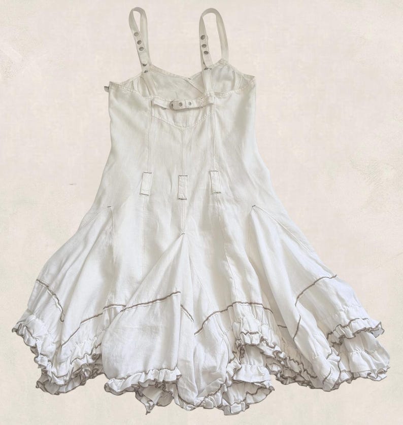K&ouml;nnte beinhalten: Ein wei&szlig;es &auml;rmelloses Kleid mit verstellbaren Tr&auml;gern und Metallbeschl&auml;gen, einem figurbetonten Oberteil mit einem horizontalen R&uuml;ckenriemen sowie einem ausgestellten Rock mit vertikalen N&auml;hten und zwei Stufen mit braunem Saum.