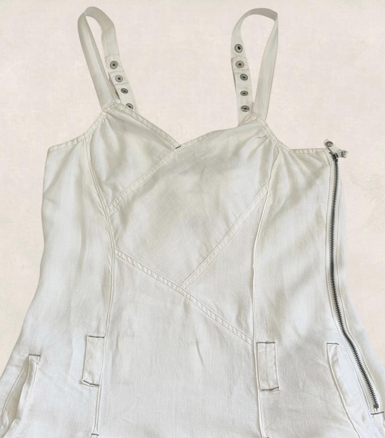 K&ouml;nnte beinhalten: Ein wei&szlig;es &auml;rmelloses Kleid mit V-Ausschnitt, breiten Schultertr&auml;gern mit silberfarbenen Metalldruckkn&ouml;pfen und einem auff&auml;lligen seitlichen Rei&szlig;verschluss. Das Kleidungsst&uuml;ck verf&uuml;gt &uuml;ber vertikale Nahtdetails und zwei kleine rechteckige Taschen.