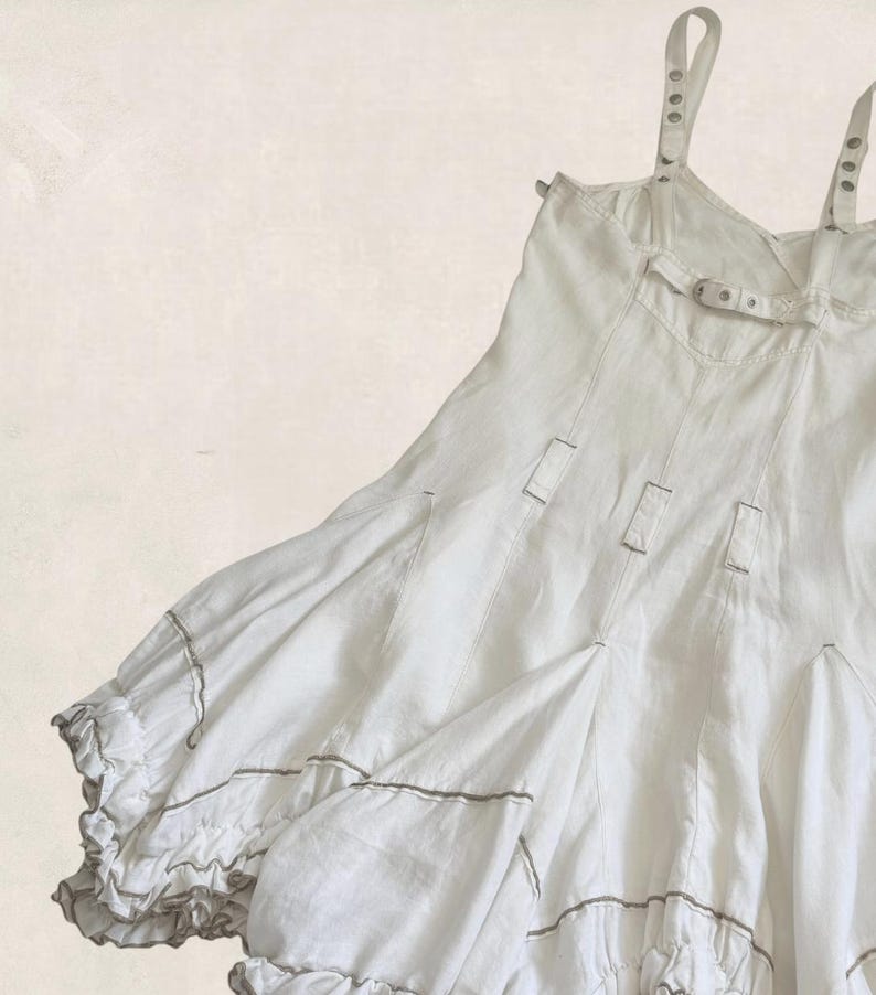 K&ouml;nnte beinhalten: Ein wei&szlig;es &auml;rmelloses Kleid mit d&uuml;nnen Schultertr&auml;gern mit Metall&ouml;sen, einem horizontalen Brustriemen mit Schnalle und vertikalen Bahnen. Der Rock hat einen volumin&ouml;sen, gerafften Saum mit ger&uuml;schten Kanten und feinen grauen Paspeldetails.