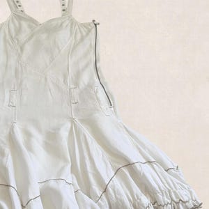 K&ouml;nnte beinhalten: Ein wei&szlig;es &auml;rmelloses Kleid mit verstellbaren Tr&auml;gern mit Metall&ouml;sen, einem seitlichen Rei&szlig;verschluss und einem ausgestellten Rock mit horizontalen Bahnen und ger&uuml;schtem Saum. Das Kleidungsst&uuml;ck ist durchgehend mit sichtbaren N&auml;hten versehen.