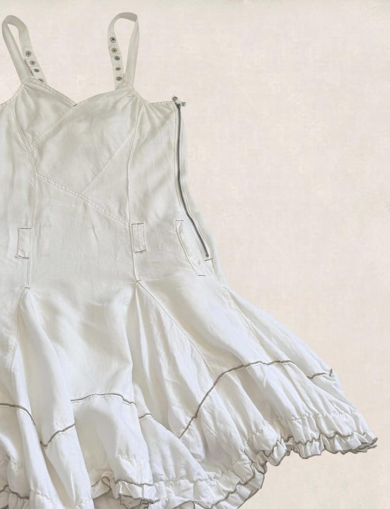 K&ouml;nnte beinhalten: Ein wei&szlig;es &auml;rmelloses Kleid mit verstellbaren Tr&auml;gern mit Metall&ouml;sen, einem seitlichen Rei&szlig;verschluss und einem ausgestellten Rock mit horizontalen Bahnen und ger&uuml;schtem Saum. Das Kleidungsst&uuml;ck ist durchgehend mit sichtbaren N&auml;hten versehen.