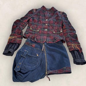 May include: A two-piece outfit featuring a plaid jacket and a denim skirt. The jacket is red, green, and black with button closures and decorative pockets. The denim skirt has a unique design with a front zipper and a draped pocket.