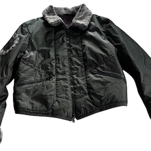 Puede incluir: Chaqueta verde oscuro con cuello y puños de piel sintética. La chaqueta tiene un diseño acolchado, cierre de cremallera y un pequeño diseño floral en la manga izquierda. La chaqueta es de estilo corto.