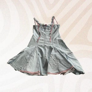 Könnte beinhalten: Hellblaues Jeanskleid mit ausgestelltem Rock und Spaghettiträgern. Das Kleid hat zwei kleine Taschen und rosa Besatz am Saum und an den Trägern. Der Rücken hat rosa Schnürdetails. Ein lässiges Kleidungsstück im Vintage-Stil.