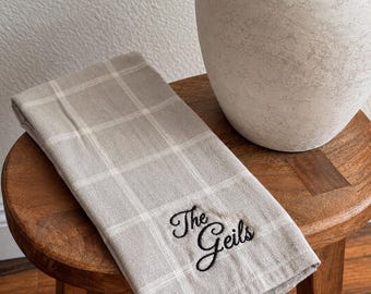 May include: A folded light gray and white plaid kitchen towel featuring the embroidered black text "The Geils" in a cursive script font. The towel is made of a woven fabric and displays a clean, grid-like pattern across its entire surface area.