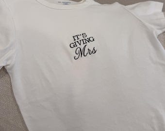 May include: A white short-sleeved t-shirt featuring black embroidered text centered on the chest. The text reads IT'S GIVING in a bold sans-serif font, with the word Mrs written below in a cursive script. The shirt has a classic crew neckline.