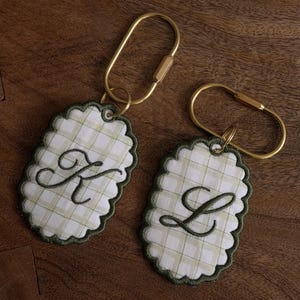 May include: Two oval-shaped keychains featuring a white and light green plaid fabric with scalloped dark green edges. Each keychain is embroidered with a dark green cursive letter, one with a K and the other with an L, attached to a gold-toned metal ring.