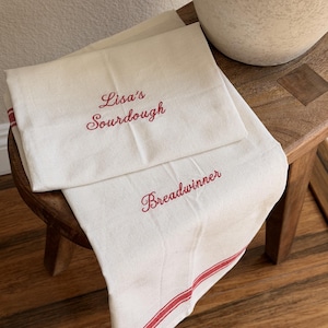May include: Two off-white kitchen towels featuring red embroidered text and red striped borders. One towel displays the text "Lisa's Sourdough" in a cursive font, while the other towel features the word "Breadwinner" in the same red script.