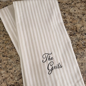 May include: A folded kitchen towel featuring a vertical striped pattern in white and light beige. The fabric is embroidered with the black cursive text "The Geils" positioned in the lower right section of the towel, showcasing a clean design.