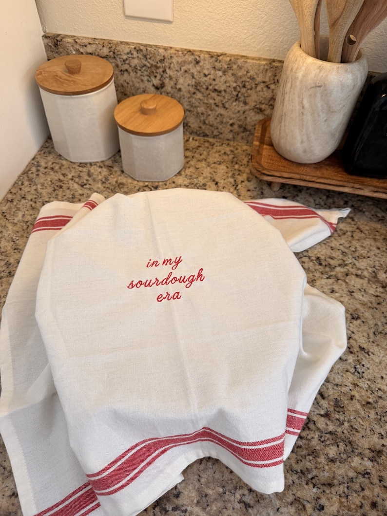 Puede incluir: Un pa&ntilde;o de cocina blanco con rayas rojas en los bordes, que presenta el texto rojo bordado "in my sourdough era" centrado en la tela. El pa&ntilde;o est&aacute; drapeado sobre un objeto redondo, mostrando una textura tejida limpia y un dise&ntilde;o sencillo.