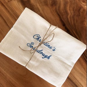 Puede incluir: Un pa&ntilde;o de tela rectangular de color blanco roto con el texto azul bordado "Christine's Sourdough" en letra cursiva. El pa&ntilde;o est&aacute; doblado y sujeto con un cordel de yute fino de color natural atado con un nudo sencillo en el centro.