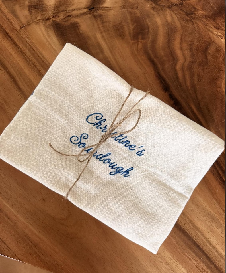 Puede incluir: Un pa&ntilde;o de tela rectangular de color blanco roto con el texto azul bordado "Christine's Sourdough" en letra cursiva. El pa&ntilde;o est&aacute; doblado y sujeto con un cordel de yute fino de color natural atado con un nudo sencillo en el centro.