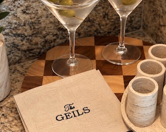 May include: A square beige linen napkin embroidered with the black text The GEILS, placed on a wooden checkerboard tray alongside two martini glasses containing clear liquid and green olives on gold picks, and several cylindrical stone cups.