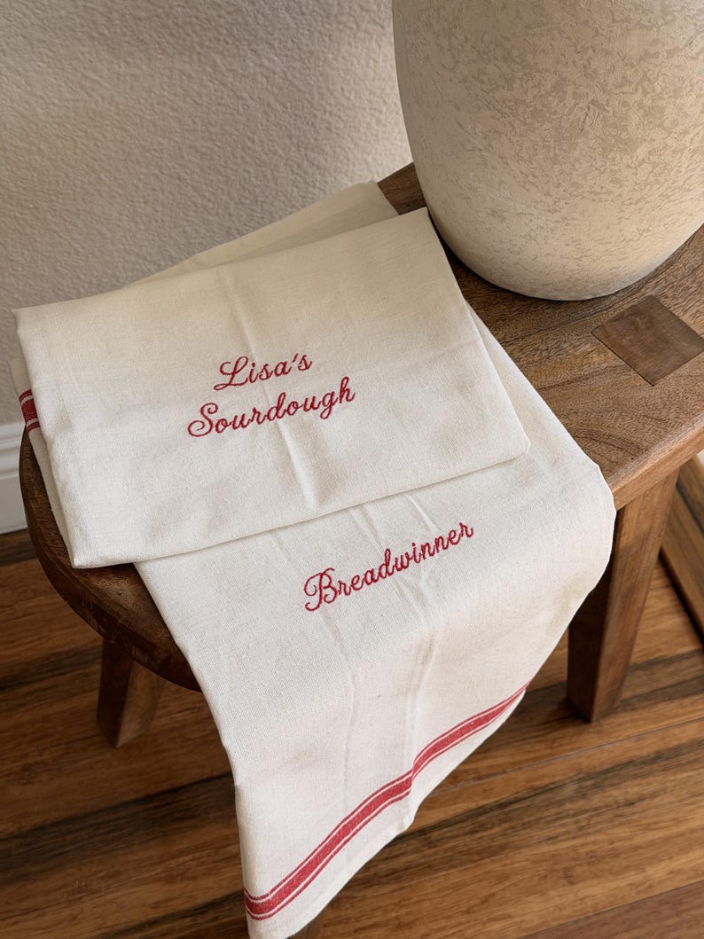 Puede incluir: Dos pa&ntilde;os de cocina de color blanco roto con texto bordado en rojo y bordes a rayas rojas. Un pa&ntilde;o muestra el texto "Lisa's Sourdough" en letra cursiva, mientras que el otro pa&ntilde;o presenta la palabra "Breadwinner" en la misma caligraf&iacute;a roja.