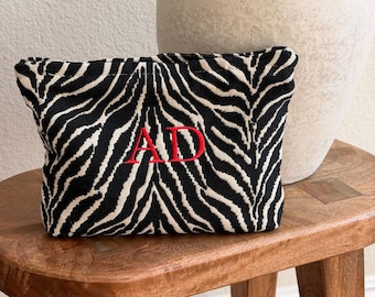 May include: A rectangular zippered pouch featuring a black and white zebra stripe pattern. The center of the pouch is embroidered with the red capital letters AD in a serif font. The fabric has a textured, woven appearance.