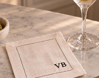 May include: A square, light beige linen cocktail napkin featuring a dark embroidered border of small dots and the monogrammed letters VB in the bottom right corner. The fabric has a visible woven texture and is placed on a flat surface.